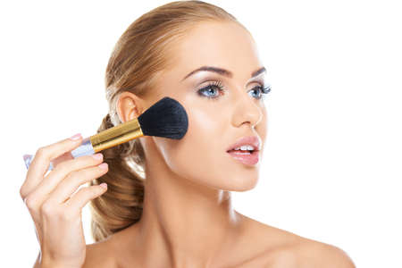 Beautiful blonde woman applying blusher or foundation powder to her cheek with a large soft cosmetics brush, beauty portrait isolated on whiteの写真素材