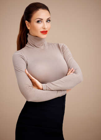 Beautiful shapely glamorous professional woman with her hair tied neatly back standing with her arms folded looking at the camera on a beige backgroundの写真素材
