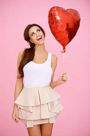 Vivacious brunette woman ina sexy miniskirt with a red heart shaped Valentine balloon in her hand on a pink studio backgroundの写真素材