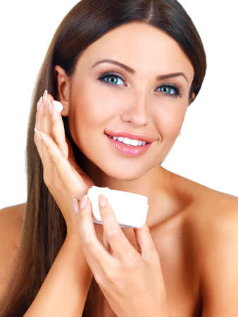 Beautiful brunette woman with a lovely smile applying face cream from a jar isolated on whiteの写真素材