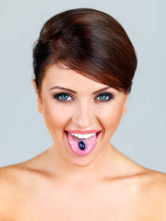 Beautiful woman with bare shoulders sticking out her tongue to show a a vitamin supplement in the form of a capsule on a grey studio backgroundの写真素材