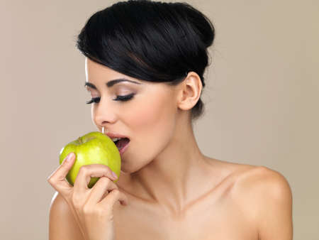 Beautiful brunette woman with bare shoulders eating a delicious fresh juicy green apple on a beige studio backgroundの写真素材