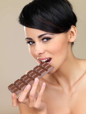 Head and shoulders portrait of a beautiful naked woman biting into a delicious bar of chocolate on a beige backgroundの写真素材