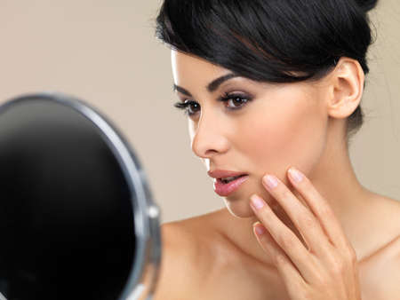 Beautiful woman looking in a hand mirror checking her complexion or makeup , cropped head and shoulders portrait on beigeの写真素材