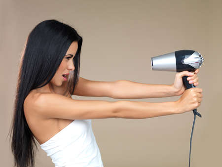 Playful woman having fun with a hairdryer holding it out in her outstretched hands pointing back at her own face isolated on beigeの写真素材
