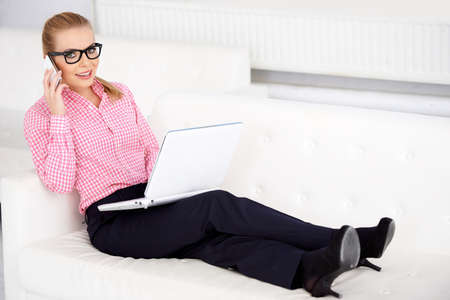 Stylish girl sitting on couch, talking on cellphone and laptop.の写真素材