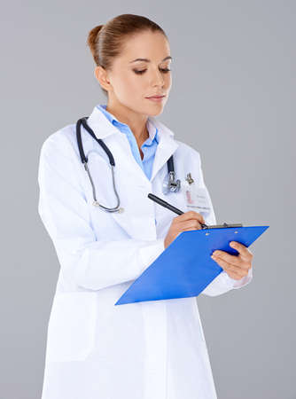 Attractive dedicated female doctor writing on a patients file taking notes on a clipboard  isolated on greyの写真素材