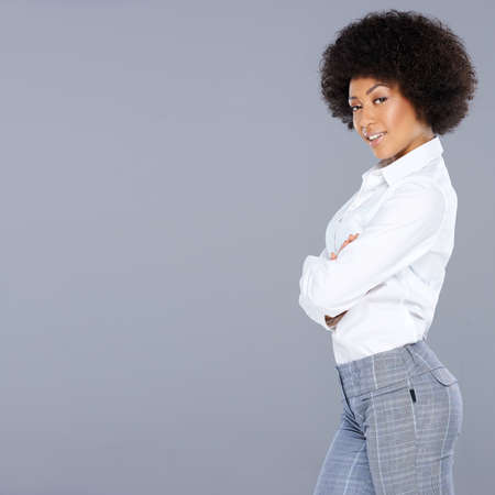 Beautiful African American woman with an afro hairstyle posing sideways with folded arms and a confident teasing expression, on grey with copyspaceの写真素材