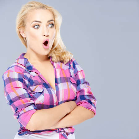 Shocked beautiful young blond woman standing with folded arms staring open mouthed towards the right of the frame on grey with copyspaceの写真素材