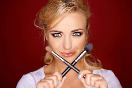 Smiling playful beautiful blond woman holding up two crossed soft cosmetic brushes in front of her face in a beauty conceptの写真素材