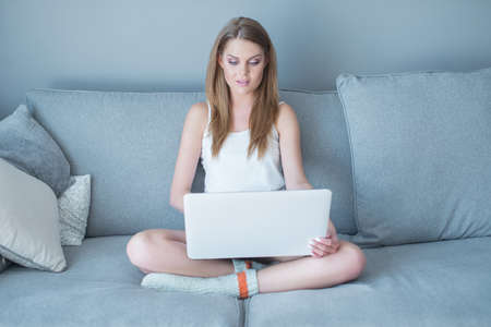 Young girl sitting cross legged on a sofa working on a laptop computer as she studies at home in comfortの写真素材