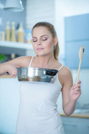 Young woman savoring the smell of her cooking holding up a stainless steel pot in the air smelling the contents with a look of bliss and satisfactionの写真素材