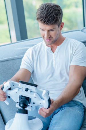Middle Age Caucasian Man in White Shirt and Faded Blue Pants Playing His Cool Gadget at Home.の写真素材