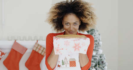 Happy young African woman holding a Christmas pastry in her hands with a smile of pleasure as she stands in front of the decorated fireplaceの写真素材