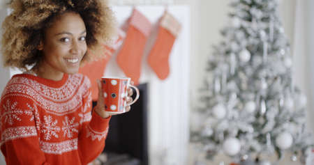 Pretty smiling young African woman in a festive red sweater drinking a mug of Christmas coffee in front of the Christmas tree at homeの写真素材