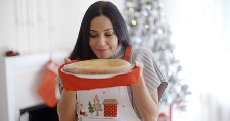 Attractive young woman baking tarts for Xmas standing in front of the Christmas tree savoring the aroma of a freshly baked pieの写真素材