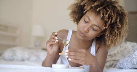 shot of a african american young woman having breakfast while lying on her bed at homeの写真素材