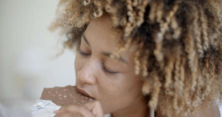 Cute young african-american woman eating chocolate in bed at homeの写真素材