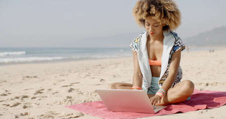 African-american woman sits on a towel in the sand with laptop on the summer beachの写真素材