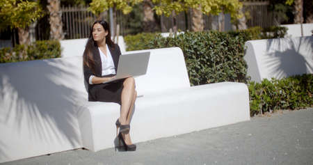 Young businesswoman using a laptop on a white park bench as she sits in the sunshine looking expectantly to the left of the frame as she waits for someone.の写真素材