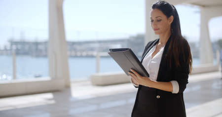 Attractive businesswoman standing on a waterfront promenade working on a tablet computer as she navigates the screen  profile viewの写真素材