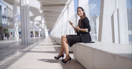 Elegant businesswoman talking on her mobile phone as she sits on a white bench in an urban street   low angle profile viewの写真素材
