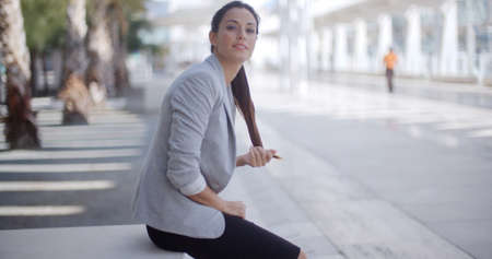 Pretty businesswoman sitting alongside a pedestrian walkway holding her long ponytail in her hand as she turns to smile at the cameraの写真素材