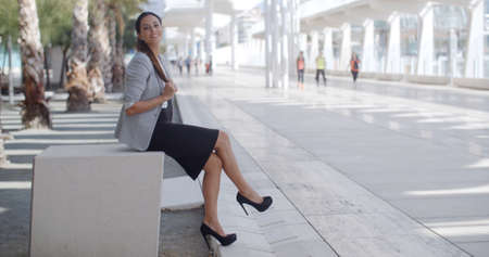 Elegant gorgeous young business woman sitting on a bench on a high key white modern urban promenade holding her long hair and smiling at the camera  with copyspace.の写真素材