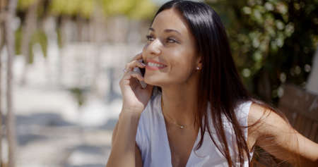 Vivacious gorgeous young woman chatting on her mobile phone with a smile of pleasure as she stands outdoors in an urban parkの写真素材