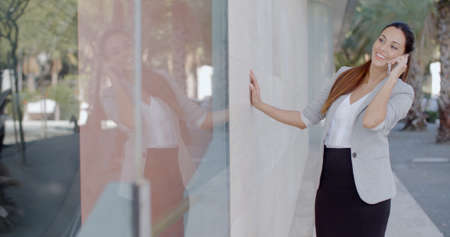 Happy relaxed young woman chatting on her mobile in an urban street as she stands with her hand resting on the wall of a commercial building.の写真素材