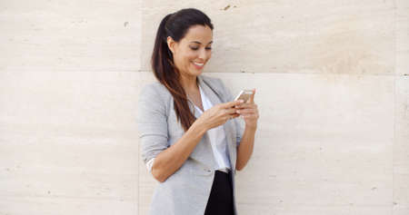 Pretty young woman standing leaning against a neutral cream colored wall with copyspace on either side checking her mobile phone for messagesの写真素材
