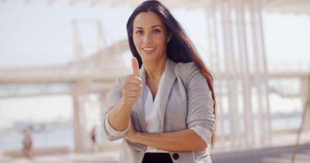 Motivated young businesswoman giving a thumbs up gesture of success and approval as she stands outdoors on a waterfront promenadeの写真素材