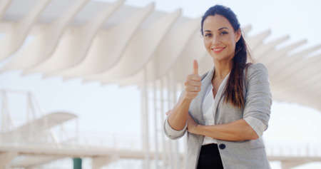 Motivated young businesswoman giving a thumbs up gesture of success and approval as she stands outdoors on a waterfront promenadeの写真素材