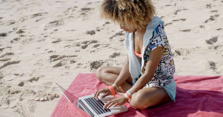 African-american woman sits on a towel in the sand with laptop on the summer beachの写真素材