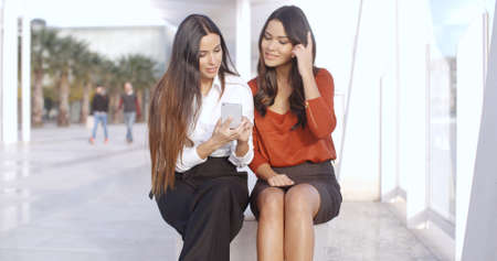 Two stylish young woman sitting outdoors in an urban esplanade checking a text message on a mobile...
