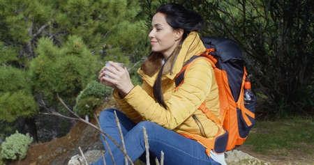 Young woman backpacker enjoying coffee as she sits cross-legged on the ground with a smile of bliss