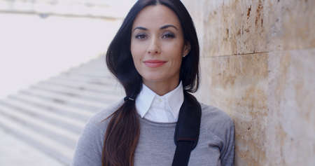 Confident young woman with gray sweater and buttoned collar leaning against marble wall past...