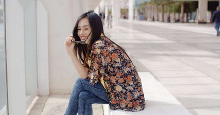 Happy vivacious young woman sitting on a bench in the sunshine in town smiling as she lowers her...