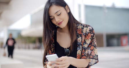 Young woman sitting reading a text message on her mobile phone on a bench at the side of an urban...