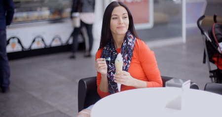 Pretty young woman sitting at a table eating an ice cream in an ice-cream parlor   shopping mall or cafeの写真素材