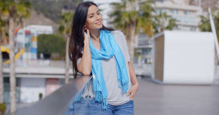 Gorgeous young grinning woman in long black hair and blue scarf with hand in hair as she leans on glass railing overlooking parkの写真素材