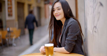 Friendly young woman sitting enjoying a beer at a table at an outdoor bar or cafeteria leaning against the wall smiling at the cameraの写真素材