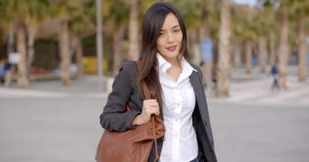 Fashionable young woman carrying a large handbag standing smiling at the camera outdoors against an...