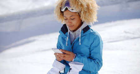 Single woman in blue snowsuit and white gloves with blond hair looking down at a phone in her hands with snowy hill in backgroundの写真素材