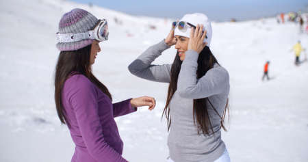Two stylish young woman chatting at a ski resort standing in a snow landscape with tourists in the distance  close up upper bodyの写真素材