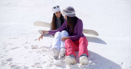 Two young women sitting side by side chatting and laughing in the snow at an alpine ski resort smiling as they relax together in the sunshine.の写真素材