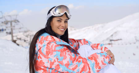 Gorgeous young brunette woman in ski clothes and goggles sitting in the snow at a mountain ski resort looking at the camera with a lovely smile.の写真素材
