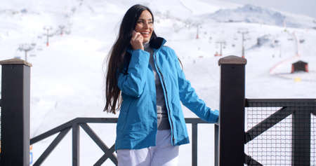 Attractive young woman at a mountain ski resort standing on a balcony overlooking the snow chatting...