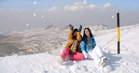 Two young women having fun in winter snow as they sit at the edge of a ski run at a mountain resort...