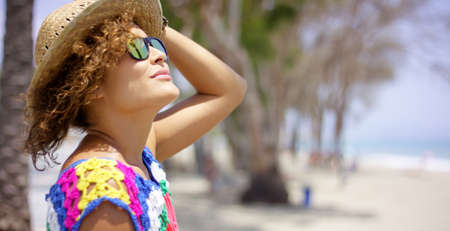 Pretty adult female in reflective yellow sunglasses and straw hat outside on beach looking upward with copy spaceの写真素材